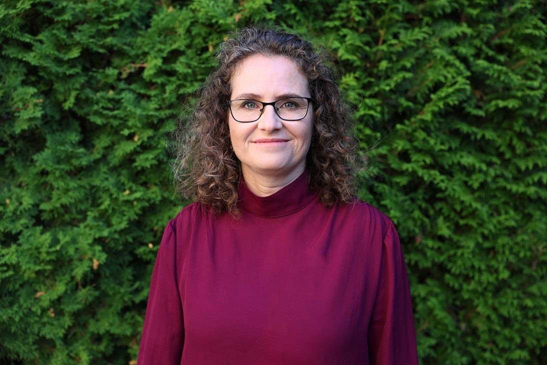 Frau mit lockigem Haar und Brille vor einer grünen Hecke